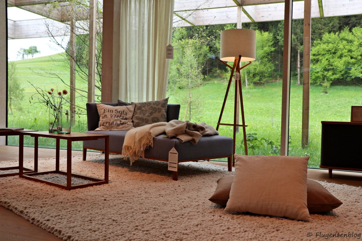graue Couch mit pastellfarbenen Polster und Decken, Stehlampe, zwei Polster liegen am Boden, heller Teppich, eckiger Couchtisch mit Blumenvase