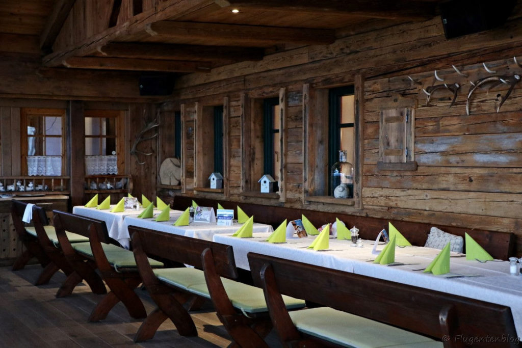 Holzvertafelung an der Wand, Holzbaenke und Holztische mit weißem Tischtuch und gruenen Servietten gedeckt