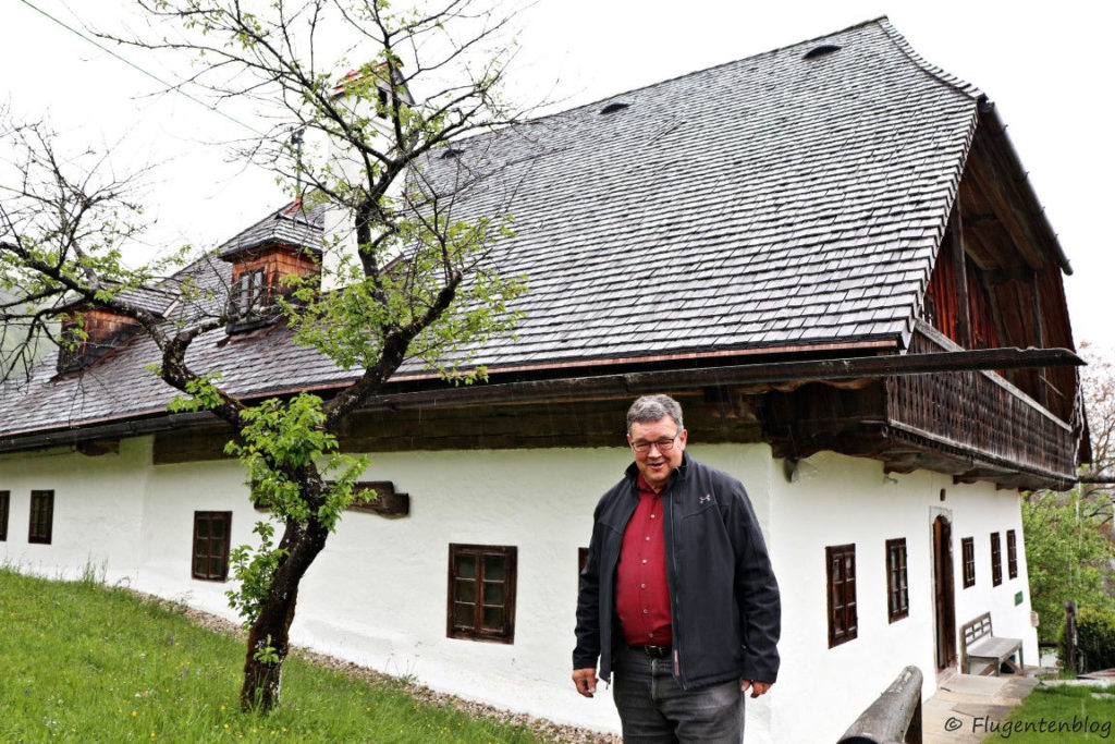 großer staatlicher Mann mit dunklen Haaren und Brille vor weißem Haus mit dunklem Dach und dunkelbraunen Holzfenstern