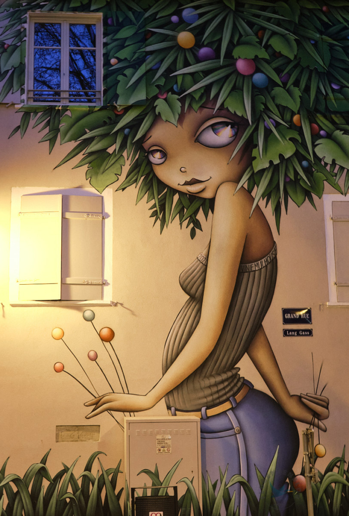 Mulhouse Street Art Mural mit Mädchen mit großen Augen, grauem Stricktop, gelben Gürtel und jeans sowie Haaren aus Blättern und bunten Kugeln