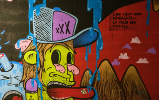 Mulhouse Graffitipolis bunter Cartoon von Mann mit gelbem Gesucht, türkis-violetter Baseballkappe, herausgestreckter Zunge mit Pflaster