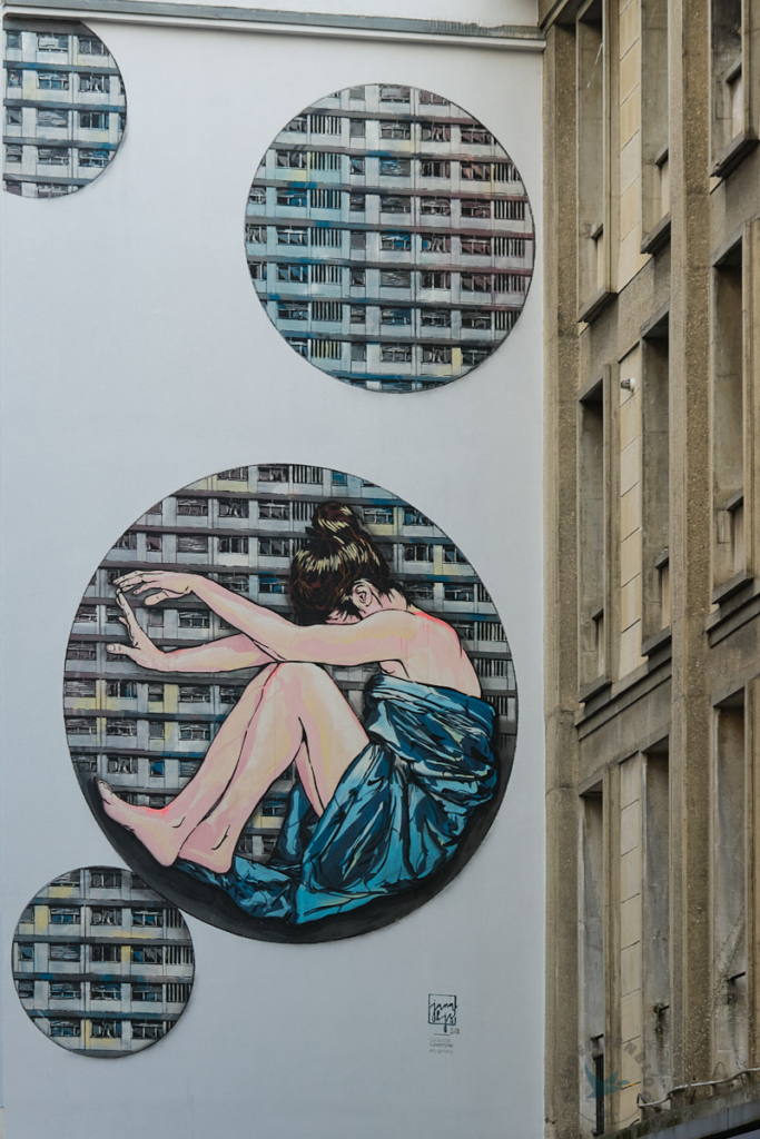 Mulhouse Streetart weiße Fassade mit runden Bildern die Häuser enthalten sowie eine sitzende Frau mit braunen hochgesteckten Haaren und blauem Kleid