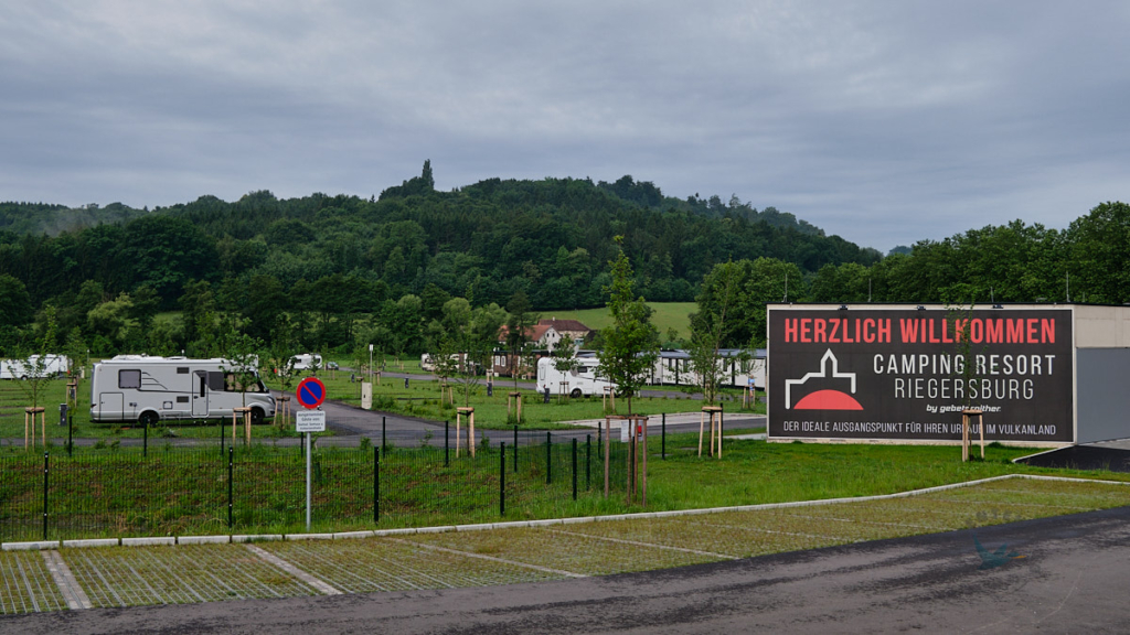 vorne Parkplaetze, rechts Willkommenstafel des Camping Resorts sowie hinter einem Zaun Wohnmobile und Mobilheime